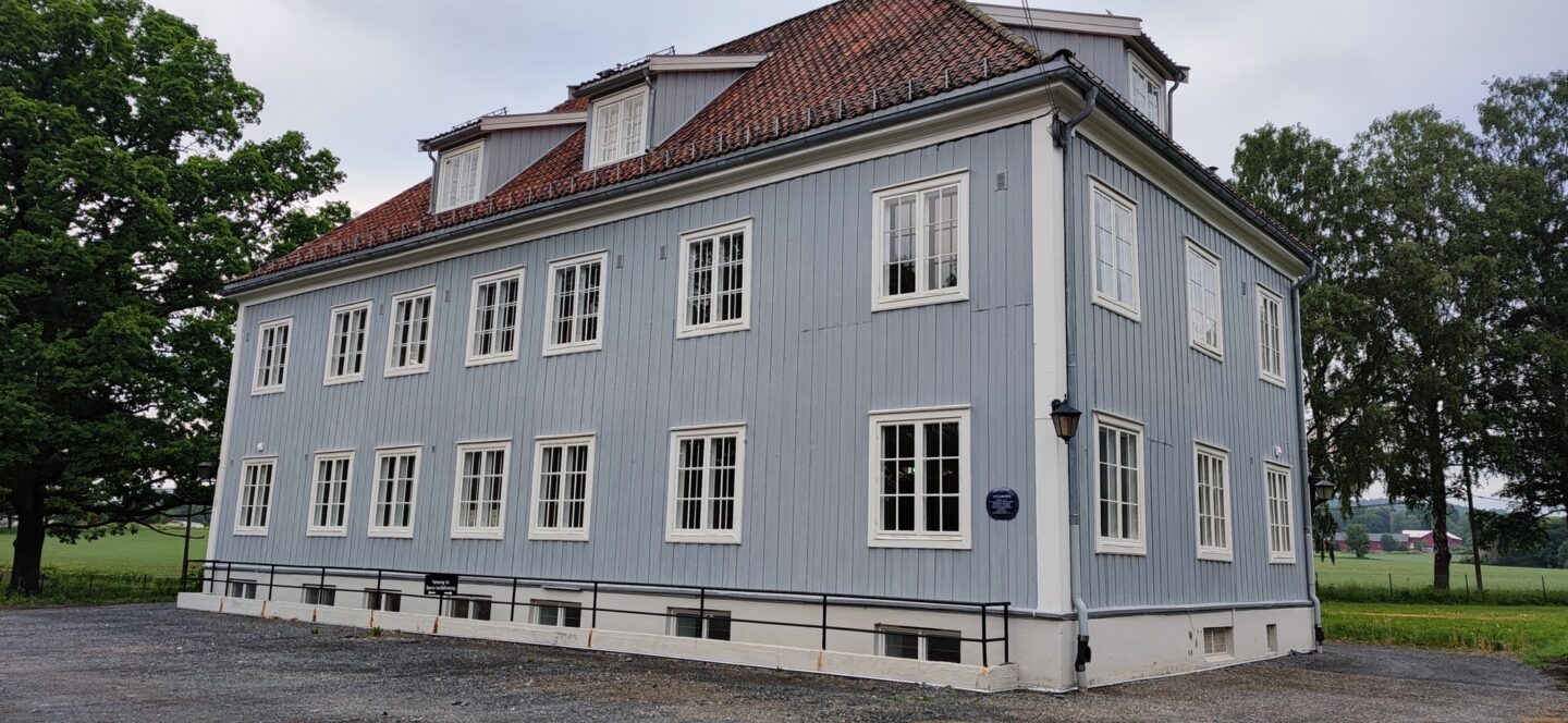 Lyst blått trehus med hvite vinduer og rødt tak, omgitt av trær og grønt landskap.